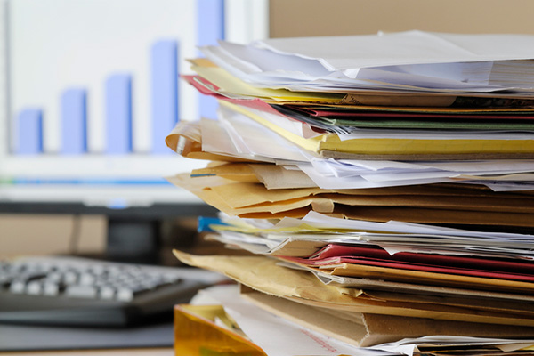 Files and papers stacked on a desk with a chart displayed in the background.