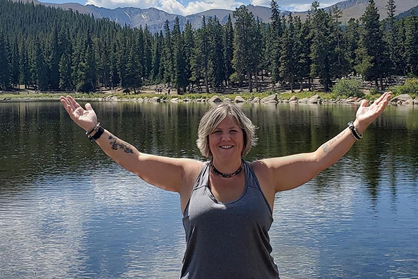 Woman stands with arms outstretched in front of a lake, forest, and mountains.