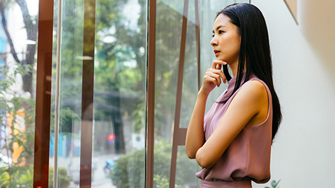 A woman standing to the side of reflective glass windows in deep thought.