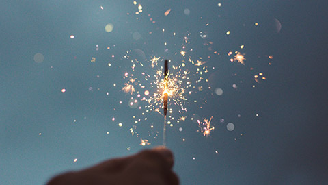 A hand holding a sparkler towards a sun-setting sky.