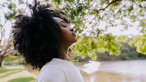 A woman standing by a lake with her eyes closed deep in thought.