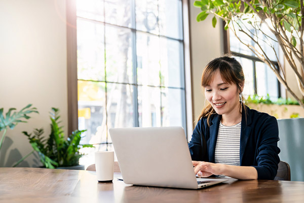 Woman works at a laptop on a table in a sunlit room.