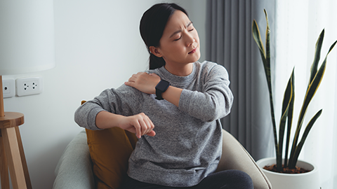 woman suffering from shoulder pain massaging on shoulder by hands sitting on armchair in living room at home.